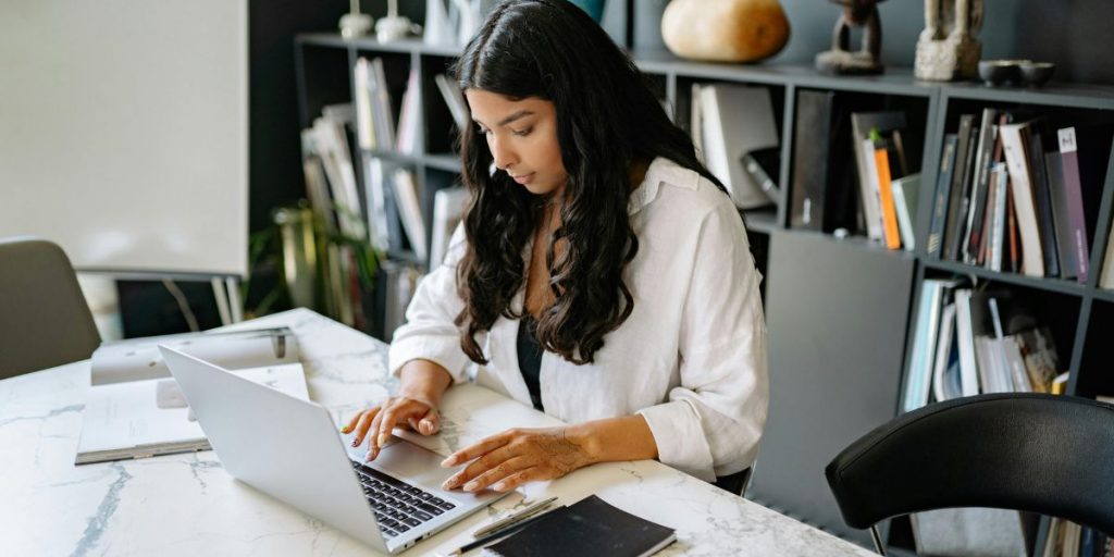 A professional woman working on a laptop in a modern office, surrounded by bookshelves and notebooks, symbolizing focus, innovation, and business preparation in a changing digital landscape.